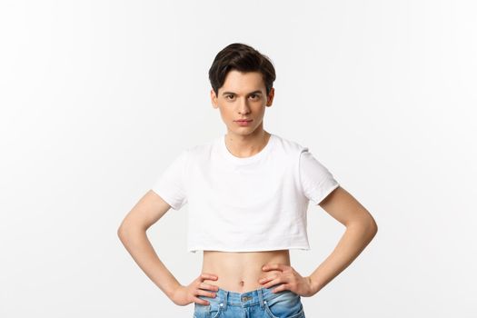Handsome androgynous man in crop top looking confident at camera, holding hands on waist. Lgbtq activist standing over white background with sassy expression.