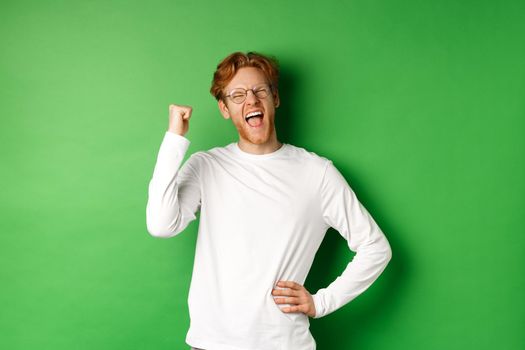 Young cheerful guy scream of joy, winning prize and celebrating, making fist pump gesture and smiling happy, standing over green background.