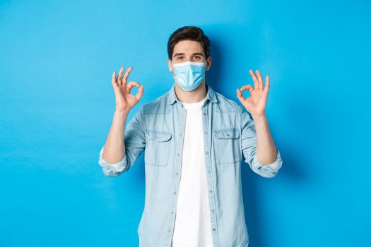 Concept of covid-19, pandemic and social distancing. Satisfied man in medical mask, showing ok signs in approval, agree or like something good, standing over blue background.