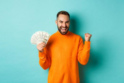 Shopping. Cheerful guy holding money, winning prize in cash and making fist pump, triumphing with satisfied expression, standing in orange sweater against turquoise background.