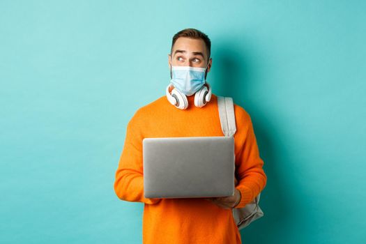 Happy young freelancer in medical mask, working on laptop, looking aside thoughtful, standing over turquoise background.