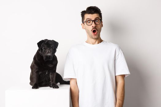 Image of dog owner and cute black pug looking at camera surprised and amazed, standing over white background.