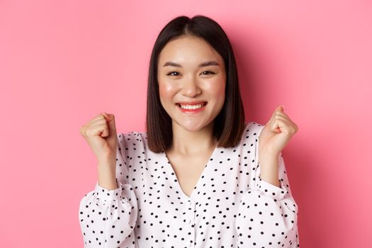 Beauty and lifestyle concept. Close-up of joyful asian woman celebrating, making fist pump and smiling of joy, winning and triumphing, standing over pink background.