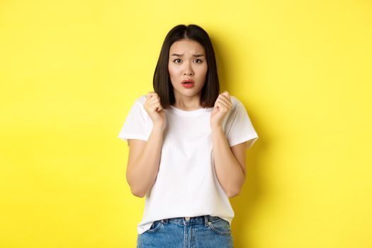 Beauty and fashion concept. Scared timid asian girl looking anxious, gasping startled and standing over yellow background.