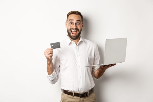Online shopping. Handsome man showing credit card and using laptop to order in internet, standing over white background.