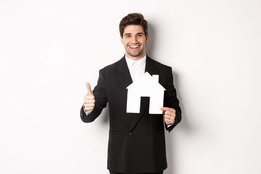 Portrait of confident real estate agent showing house maket and thumb-up, standing against white background.