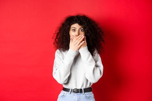 Image of speechless caucasian woman covering mouth with hands, looking shocked at camera, standing in casual clothes on red background.