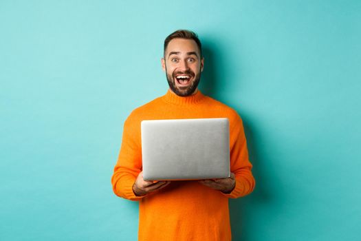Happy man using laptop and looking excited at camera, standing with computer against light blue background.