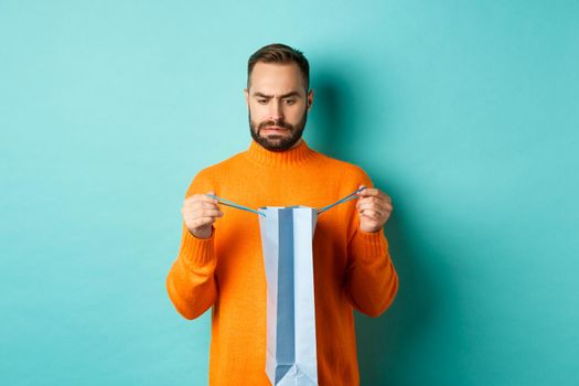 Disappointed man open shopping bag and dislike gift, frowning displeased, standing in orange sweater against turquoise background.