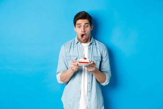 Man looking shocked at birthday cake, standing against blue background.