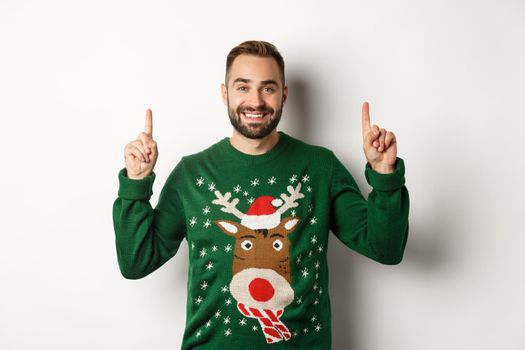 Winter holidays and christmas. Handsome bearded man in green sweater, pointing fingers up and smiling, showing advertisement, white background.