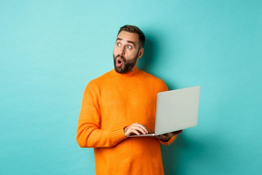 Handsome man doing online shopping, looking up thoughtful while using laptop, standing over light blue background.