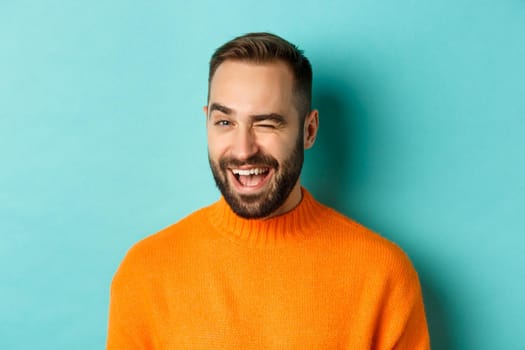 Handsome confident man winking at camera, smiling sassy, standing in orange sweater against blue background.