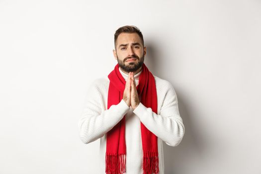 Christmas holidays and New Year concept. Desperate man begging for help, asking for favour, holding hands in pray and looking with hope at camera, standing over white background.