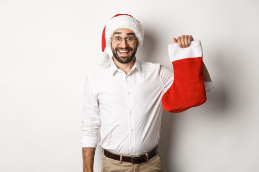 Merry christmas, holidays concept. Excited bearded guy in santa hat holding xmas sock and smiling, celebrating New Year, white background.