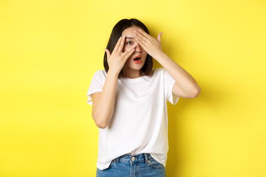 Intrigued asian girl waiting for surprise, peeking through fingers on eyes, smiling happy, standing over yellow background.