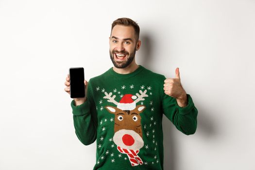 New Year, holidays and celebration. Handsome bearded man showing mobile screen and thumb up, approve something, standing over white background.