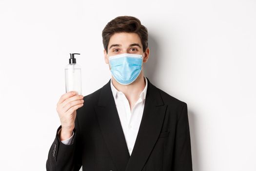 Concept of covid-19, business and social distancing. Close-up of handsome man in trendy suit and medical mask, showing hand sanitizer, standing against white background.