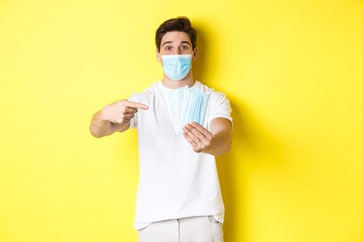 Concept of covid-19, quarantine and preventive measures. Young caucasian man giving medical masks for you, standing against yellow background.