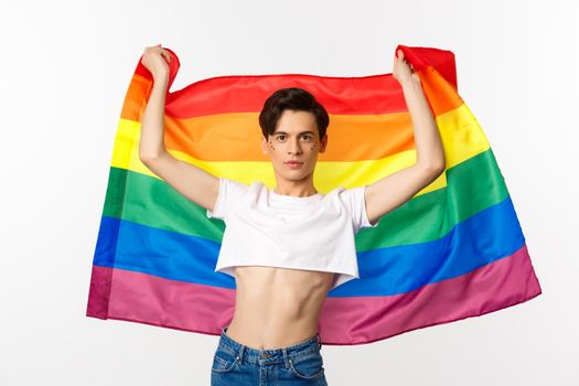 Young man in crop top, with glitter on face, raising pride flag with confident emotion. Queer person smiling with a lgbt flag.