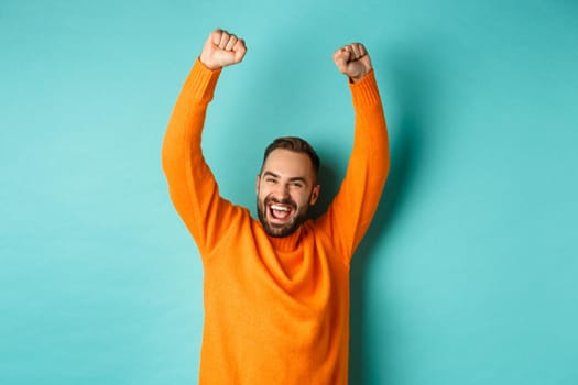 Waist-up shot of happy guy triumphing, raising hands up and rejoicing of winning, celebrating victory, standing against turquoise background.