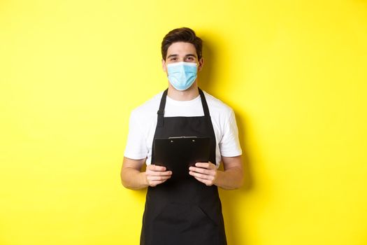 Concept of covid-19, small business and quarantine. Salesman in black apron and medical mask holding clipboard, working in shop, standing over yellow background.