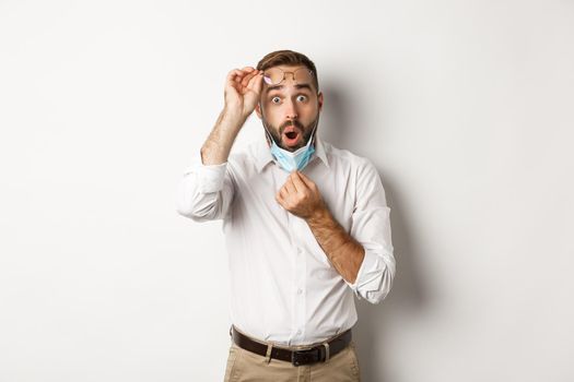 Covid-19, social distancing and quarantine concept. Surprised man take-off face mask and glasses, looking at something impressive, white background.