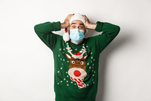Concept of covid-19 and Christmas holidays. Excited and super happy man in medical mask and Santa hat looking amazed, white background.