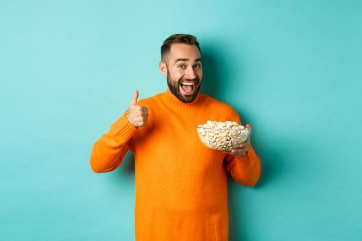 Satisfied young man, showing thumb-up and smiling, eating popcorn and watching good movie or tv, standing over blue background.