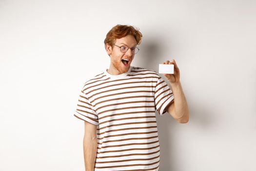 Shopping and finance concept. Excited young man with red hair, staring at plastic card and scream of joy, standing over white background.