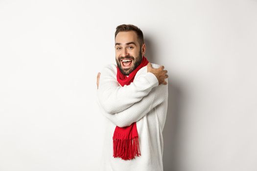 Happy man getting warm, hugging himself and feeling chilly on christmas winter holidays, wearing sweater with red scarf, standing over white background.