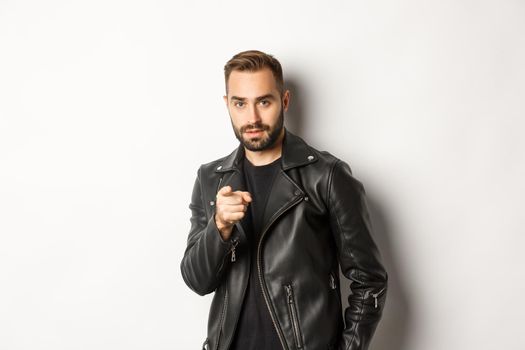 Handsome and cool bearded guy pointing finger at camera, wearing leather jacket, standing sassy against white background.