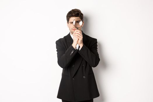 Image of serious businessman in black trendy suit, looking through magnifying glass, looking for employees, standing against white background.