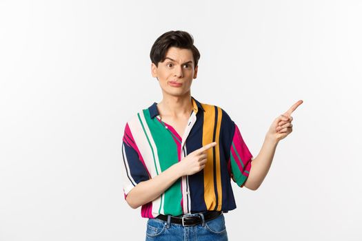 Skeptical gay man grimacing disappointed, pointing fingers right at bad product, standing over white background.