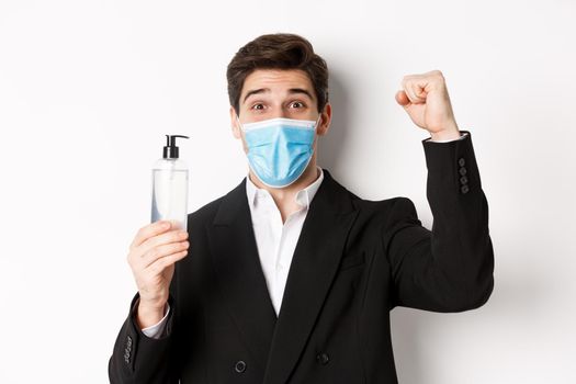 Concept of covid-19, business and social distancing. Close-up of happy man in trendy suit and medical mask, cheering and raising hand up, showing hand sanitizer, white background.