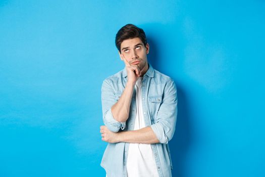 Annoyed adult man rolling eyes and looking bored, standing indifferent against blue background in casual outfit.