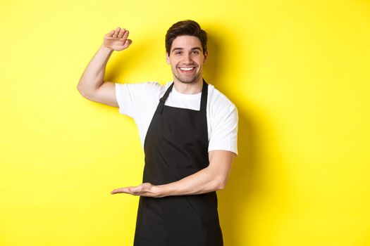 Smiling handsome barista showing something long or large, standing over yellow background.