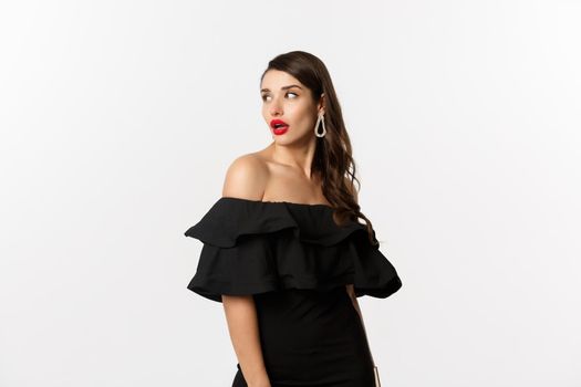 Fashion and beauty concept. Image of attractive young woman in black dress turn behind and looking at copy space, standing over white background.