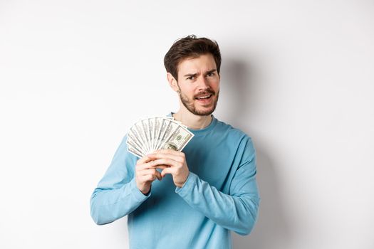 Handsome young man showing money and looking pleased. Guy dancing with dollars, earn income, standing over white background.