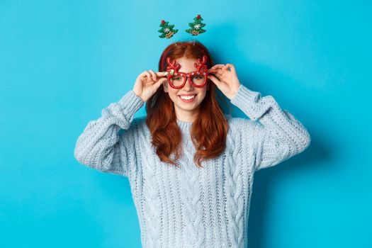 Winter holidays and Christmas sales concept. Beautiful redhead female model celebrating New Year, wearing funny party headband and glasses, smiling at camera.