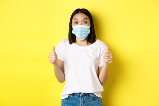 Covid-19, quarantine and social distancing concept. Cheerful asian woman in medical mask and white t-shirt showing thumbs up in approval, praise good deal, yellow background.