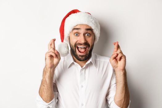 Party, winter holidays and celebration concept. Happy man in santa hat making christmas wish, cross fingers for good luck and looking excited, white background.