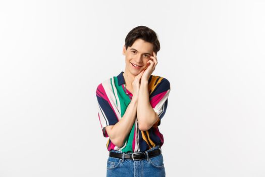 Beautiful young man looking with admiration at camera, smiling, gently touching face, standing over white background.