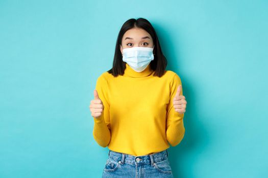 Covid-19, social distancing and pandemic concept. Excited asian woman showing thumbs up and looking impressed, praising good deal, wearing face mask, blue background.