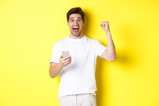 Cheerful man winning on smartphone, raising hand up and holding mobile, achieve app goal, standing over yellow background.