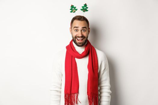 Christmas party and celebration concept. Happy bearded man looking surprised, wearing funny accessory hat, standing against white background.