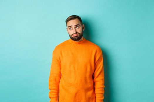 Sad and gloomy man sulking, looking bored at upper left corner, standing in orange sweater against light blue background.