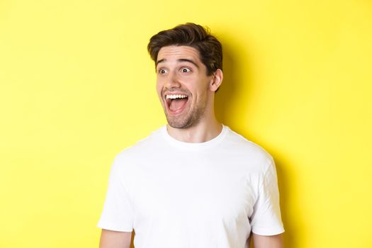 Image of happy man checking out promo, looking left with amazement, standing in white t-shirt against yellow background.