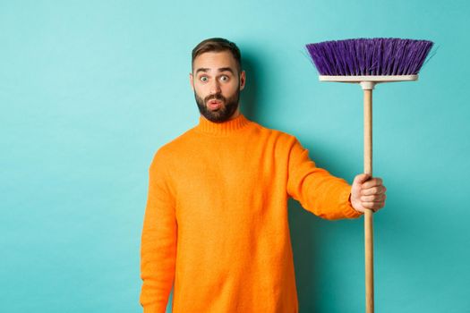 Shocked man receiving broom to do house chores, looking confused, standing over light blue background.