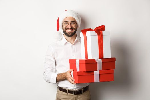 Merry christmas, holidays concept. Happy young man smiling, holding gifts in boxes and wearing santa hat, white background.